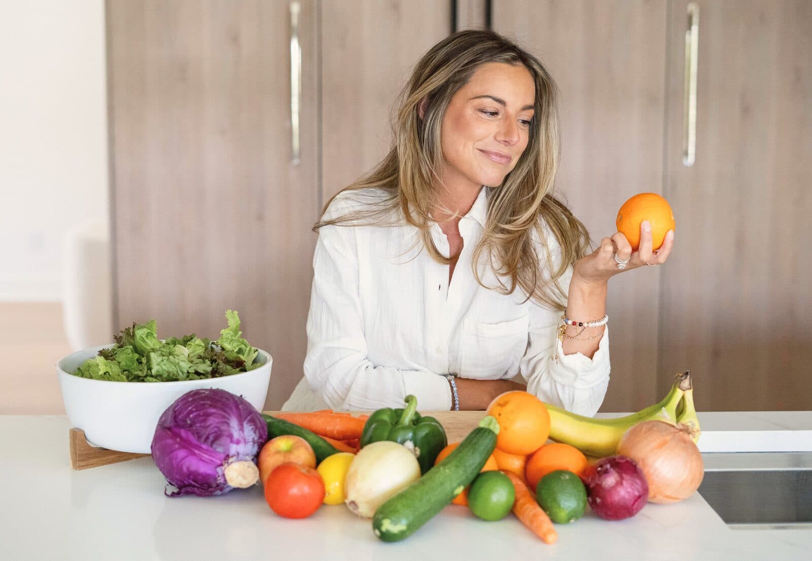 Danielle smiling at an orange in her hand, with fresh produce on the kitchen counter