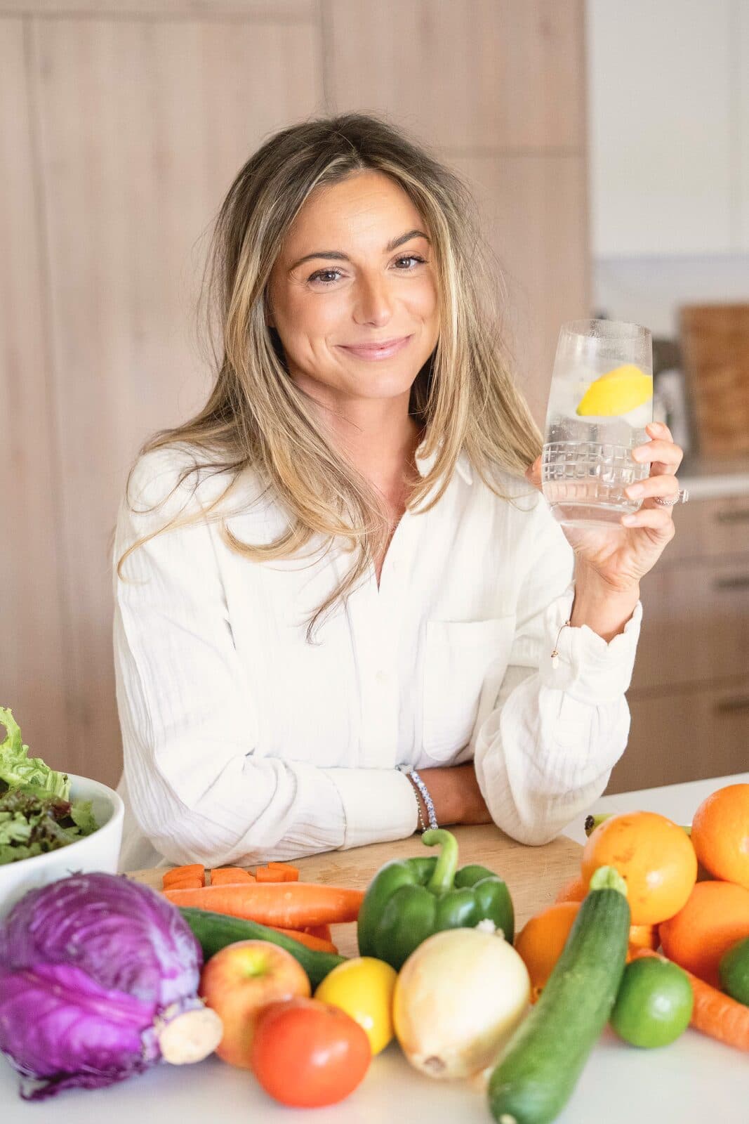 Danielle smiling at her kitchen counter with a glass of lemon water and a spread of fresh vegetables and fruit